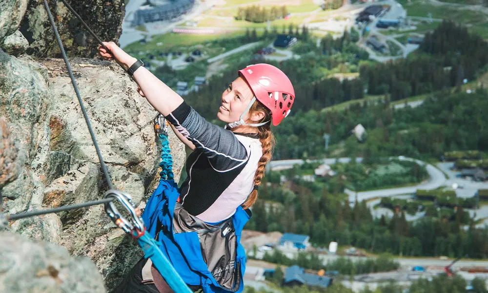 Jente klatrer via ferrata med utsikt over grønne Hemsedal langt der nede