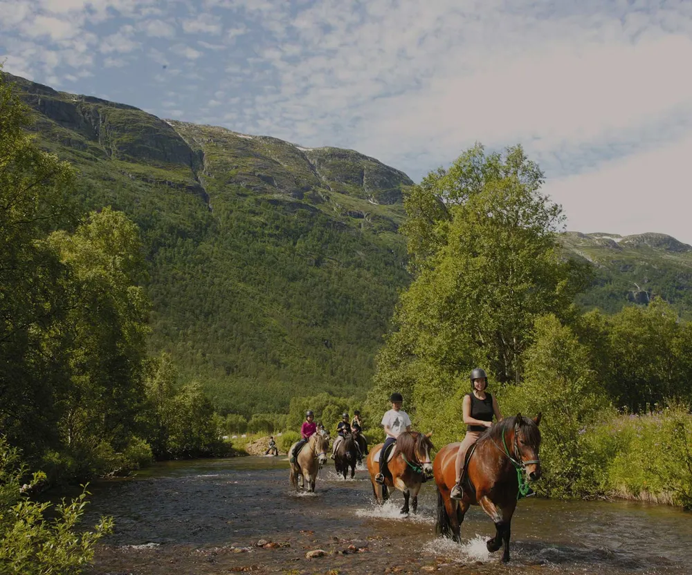 Rideturfølge krysser elva i en frodig grønn fjelldal i Hemsedal