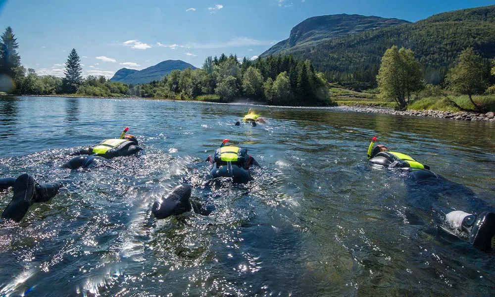 Gruppe i våtdrakter snorkler nedover en klar fjellelv i sol